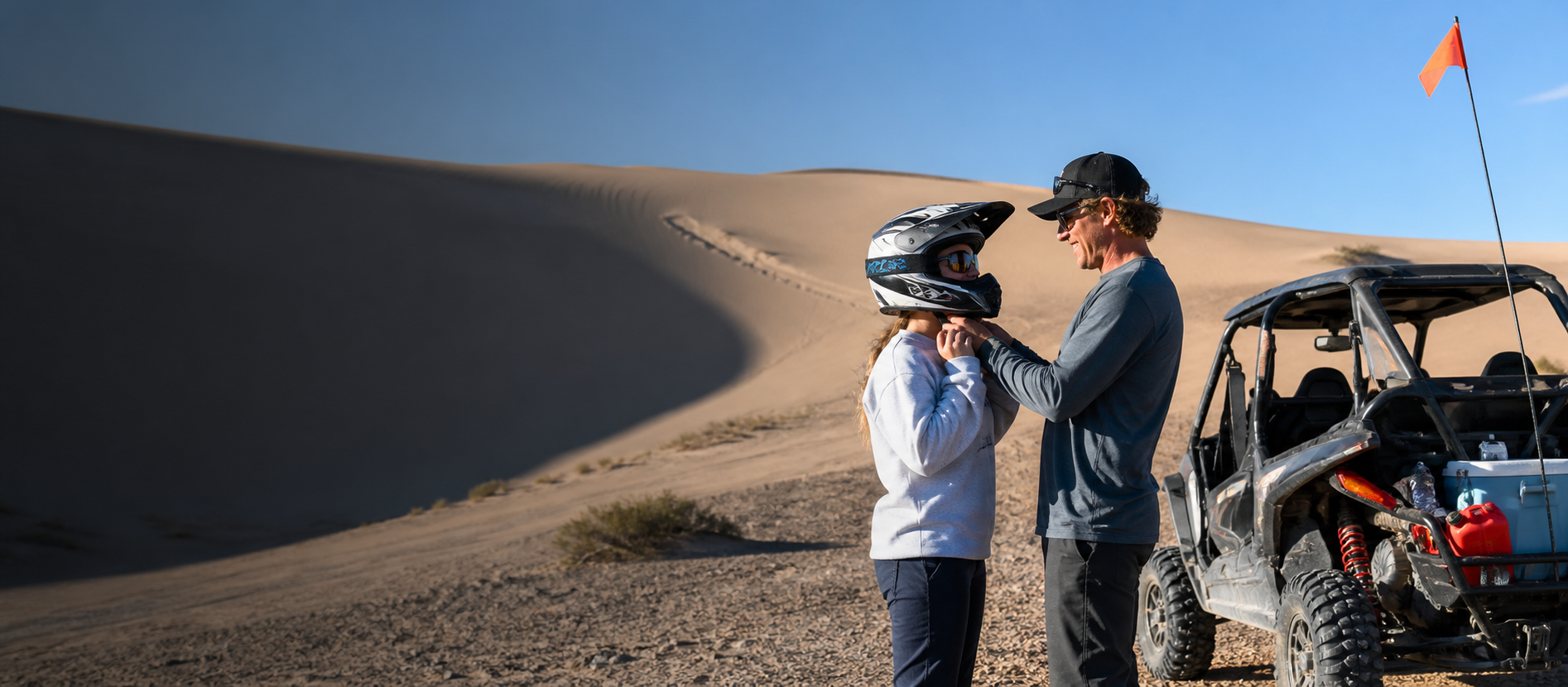 An experienced Sand Mountain rider helping a first-time visitor secure a helmet beside a flagged side-by-side