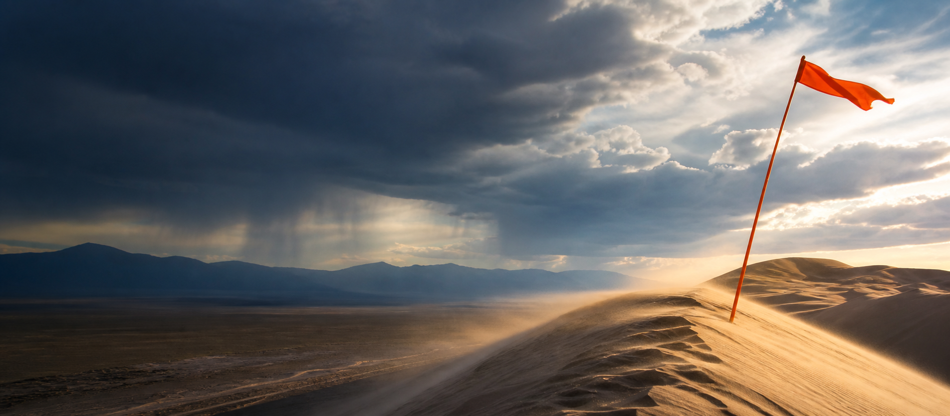 A changing high-desert weather front with rain, blowing sand, and an orange safety flag over Sand Mountain